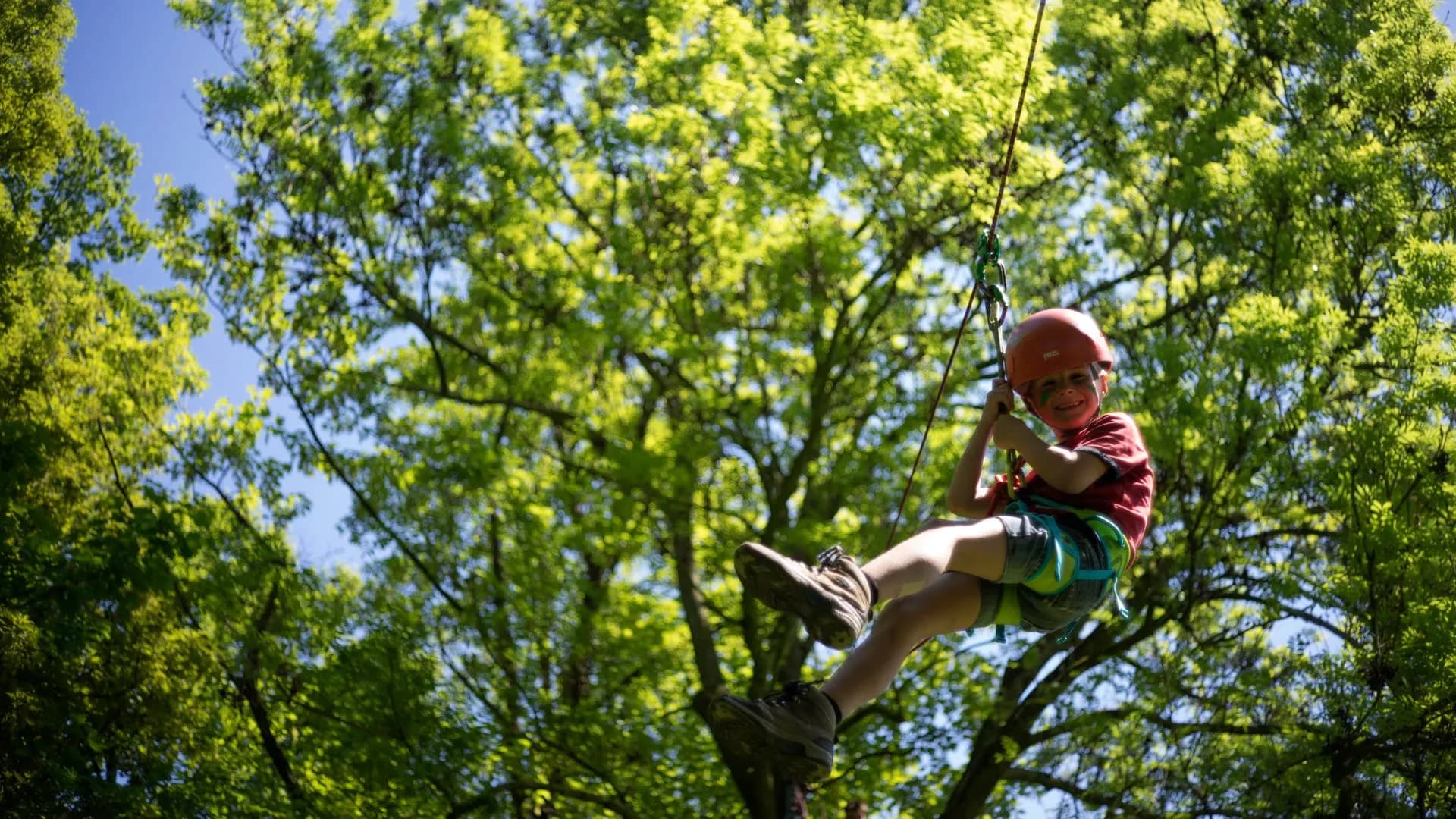 Tokkelbaan van een hoogteparcours in actie, met een kind veilig vastgemaakt aan een kabel, glijdend door een groene, bosrijke omgeving.
